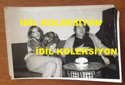 TANJU OKAN'IN ORİJİNAL FOTOĞRAFI - 18 x 12 cm EBADINDA - TANJU OKAN'IN EŞİ ZERRİN OKAN ve KÖPEKLERİYLE BİRLİKTE EVLERİNDE ÇEKİLMİŞ ÇOK GÜZEL BİR FOTOĞRAF (ZERRİN ERDOĞAN, TANJU OKAN'IN İKİNCİ EŞİDİR. AİLESİNİN TÜM ENGELLEMELERİNE RAĞMEN EVLENİRLER, ANCAK DAHA SONRA AİLE NE YAPIP EDİP ZERRİN'İ KÖŞKE KAPATIR ve TANJU OKAN KÖŞKÜN GİRİŞİNDE GÜNLERCE BEKLER, AİLE YİNE DE ZERRİN'İ GİZLİCE AMERİKA'YA YOLLAR ve BU BİRLİKTELİĞİ SONLANDIRIR. TANJU OKAN BU AYRILIĞIN SONRASINDA KENDİNİ YENİDEN ALKOLE TESLİM EDER ve ÇAREYİ SARHOŞ OLUP UNUTMAKTA ARAR...)