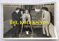 TÜRK SANAT MÜZİĞİ SANATÇISI SEVİM TUNA'NIN ORİJİNAL 2'Lİ FOTOĞRAF SETİ - 18 x 12 cm  EBADINDA - SEVİM TUNA YANINDAKİLERİN YARDIMIYLA KOLTUK DEĞNEKLERİYLE YÜRÜRKEN ÇEKİLMİŞ 2 FARKLI KARE