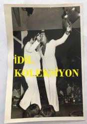 TÜRK HALK MÜZİĞİ SANATÇISI AHMET SEZGİN'İN ORİJİNAL FOTOĞRAFI - 18 x 12 cm EBADINDA - AHMET SEZGİN BEYAZ TAKIM ELBİSESİYLE YER ALDIĞI SAHNEDE TÜRKÜ OKUYUP DİNLEYİCİYLE KARŞILIKLI OYNARKEN ÇEKİLMİŞ GÜZEL BİR KARE