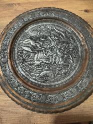 bu tabak, üzerindeki detaylı figürlerle yüksek dekoratif değere sahiptir. Hem koleksiyonluk hem de şık bir duvar aksesuarı arayanlar için kaçırılmayacak bir parçadır. Orta bölümde at üzerinde kılıçlı süvari ve av sahnesi detaylı kabartma (repousse) tekniğiyle işlenmiştir. Tabağın bordür kısmı stilize çiçek ve bitki motifleriyle bezelidir. Bakır üzerine kalay kaplama olup, zamanla oluşan doğal patini sayesinde otantik bir görünüm kazanmıştır. Arkasındaki askı halkası sayesinde duvar dekoru olarak kullanılabilir; şark köşeleri, ofisler veya salonlar için uygundur