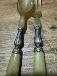 Antika Gümüş İşlemeli Boynuz Servis Takımı Zarafeti ve doğallığı bir arada sunan, koleksiyonluk servis seti (kaşık ve çatal). Özel gün sofralarınıza prestij katacak ya da dekoratif olarak sergilenecek nadide bir parçadır. Tutma yerleri ve uç kısımları doğal boynuz/kemik dokulu özel malzeme. Orta kısımları ince işçilikli, gümüş kaplama kabartma motiflerle bezelidir. Yaşına bağlı hafif bir patina (eskimiş doku) almış olup, herhangi bir kırığı bulunmamaktadır. Vintage/Antika severler için idealdir. Salata servisi veya dekoratif amaçlı uygundur.