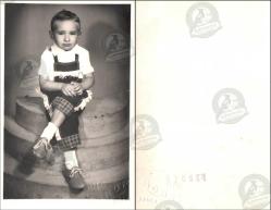 Foto Rekor, Adana, Fotoğraf Stüdyosu, Fotoğraf, 1950’ler: Basamaklı platform üzerinde oturan küçük bir çocuk, önlük ve kısa pantolonla ciddi ifadeli portre pozu.