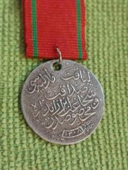 Kılıçlı Gümüş Liyakat Madalyası.. H.1332 M. 1916 Tarihli Gümüş Kılıçlı APARATI Olan Koleksiyonluk Liyakat Madalyası..  1890 tarihli Osmanlı dönemi Liyakat Madalyası. Üzerinde Osmanlı tuğrası, silah ve sancak motiflerinden oluşan devlet arması bulunan bu madalya, arka yüzünde Osmanlıca hat ile yazılmış metni ve altında 1308 (1890/1891) tarihini taşımaktadır. Osmanlı Devleti'nde liyakat ve hizmet karşılığı verilen bu madalyalar, devlet görevlileri ve askerî hizmet sahiplerine takdim edilirdi. Tarihi ve koleksiyon değeri yüksek olan bu eser, Osmanlı nişan ve madalya koleksiyonları için dikkat çekici nadir örneklerden biridir.