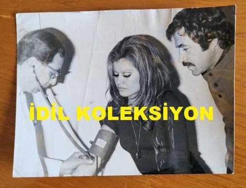 OYUNCU ve SES SANATÇISI SERPİL ÖRÜMCER'İN ORİJİNAL FOTOĞRAFI - 15,5 x 12 cm EBADINDA - SERPİL ÖRÜMCER, ŞARKICI BERKANT'LA BİRLİKTE TANSİYON ÖLÇÜMÜ YAPTIRIRKEN ÇEKİLMİŞ BİR KARE